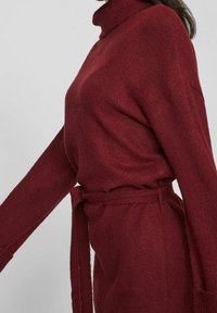 Robe-pull en tricot bordeaux avec col montant, manches longues et ceinture nouée en tissu à la taille. Texture douce et coupe décontractée.