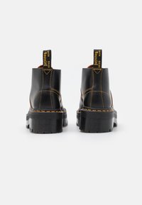 Botas de tobillo de cuero negro con una suela de plataforma gruesa, costuras amarillas y una tira de agarre con el texto "CON SUELAS RESBALOSAS".
