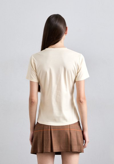 Cream-colored short-sleeve t-shirt with a round neckline, fitted shape, and smooth texture, paired with a brown plaid pleated skirt.