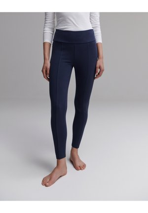 Kobieta w granatowych legginsach z wysokim stanem i przednimi szwami, stojąca boso na szarej podłodze na tle szarej ściany.