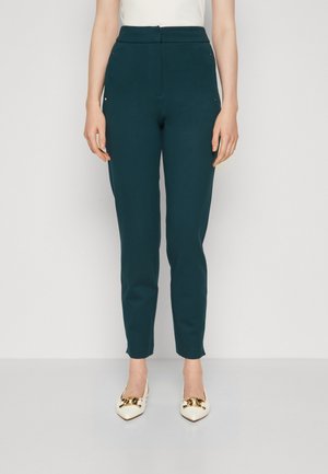 Kobieta nosząca ciemnozielone spodnie wysoki stan, zwężane oraz białe, szpiczaste baleriny ze złotym łańcuszkiem, stojąca na jednolitym tle.