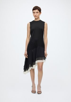 Γυναίκα με μαύρο αμάνικο φόρεμα με ασύμμετρο στρώσιμο στο τελείωμα σε μαύρο και άσπρο, συνδυασμένο με μαύρες ανοιχτές γόβες.