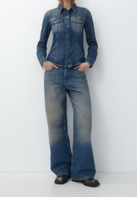 Chaqueta de denim con un diseño ajustado, color azul desteñido y bolsillos frontales, combinada con jeans de pierna ancha en azul con un degradado más claro en el dobladillo.