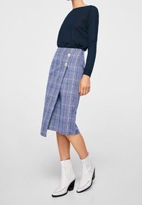 Kobieta w granatowej bluzce z długim rękawem, niebieskiej spódnicy w kratę z dwiema guzikami oraz białych botkach na kostkę z blokowym obcasem, stojąca na szarym podłożu.