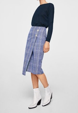 Femme portant un haut à manches longues bleu marine, une jupe en tartan bleue à enrouler avec deux boutons, et des bottines blanches à talons block, se tenant sur un sol gris.