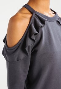 Sudadera de color gris oscuro con diseño de hombro descubierto y acentos con volantes a lo largo del escote y los bordes de las mangas. Textura suave con ribete acanalado.