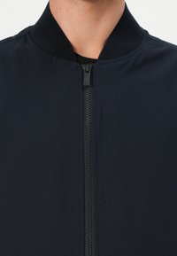 Jachetă bomber neagră cu un finisaj neted, guler ribbed și un fermoar central. Prezintă un design minimalist, fără modele vizibile.