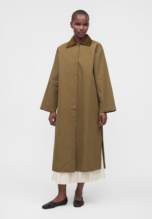 Model draagt een lange olijfgroene jas met brede mouwen en een bruine kraag over een witte rok en zwarte ballerina's, staand op een witte achtergrond.