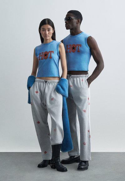 Top cropped azul com estampa "HOT", calças de fato de treino cinzentas com estampas de lábios vermelhos, acessório felpudo azul e botas pretas. Fundo simples.