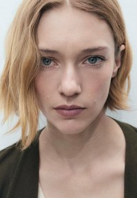 Detailný záber osoby s krátkymi vlnitými blond vlasmi a svetlomodrými očami. Majú neutrálny výraz a sú oblečení v tmavozelenom oblečení.