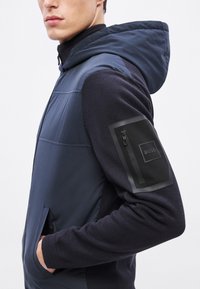 Nodilumizēts tumši zils vestes ar kapuci; raksturo gluds auduma ķermenis un ribots piedurknes. Iekļauta kabata ar rāvējslēdzēju uz kreisā rokas ar logo plāksteri.