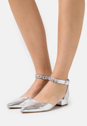 Chaussures à talon bas argentées à bout pointu avec des brides de cheville embellies de cristaux, portées pieds nus sur fond blanc.