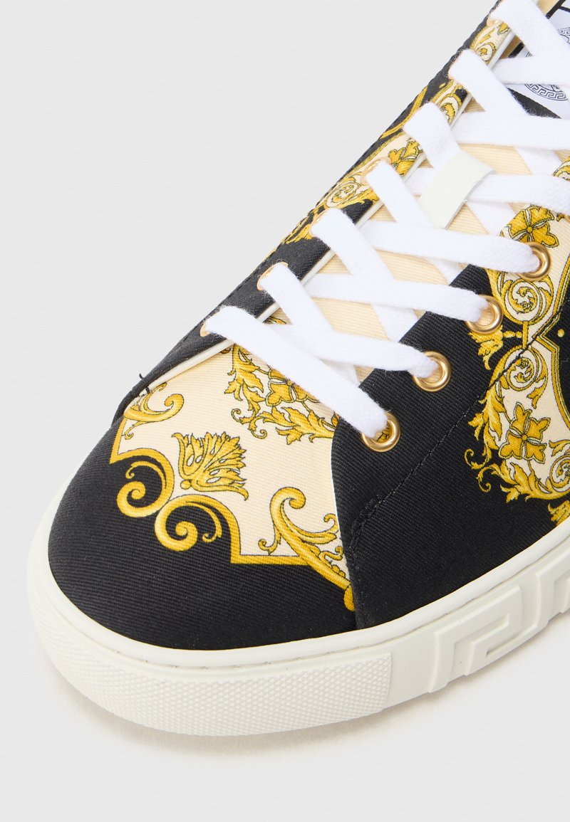 Schwarz-gold gemusterter Sneaker mit einem Obermaterial aus Canvas, weißen Schnürsenkeln, goldfarbenen Ösen und einer strukturierten Gummisohle.