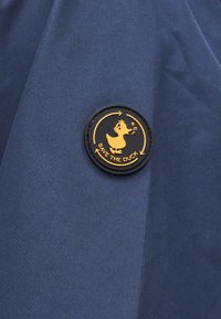 Στρογγυλό μαύρο και κίτρινο σήμα σε σκούρο μπλε ύφασμα που δείχνει σιλουέτα πάπιας που τραγουδά με βέλη και το κείμενο «SAVE THE DUCK».