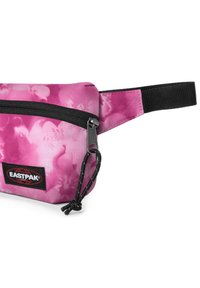 Růžová ledvinka vyrobená z látky, s hladkým povrchem, tmavým zipem a nastavovacími popruhy. Výrazná pro květinové vzory a nášivku s logem.