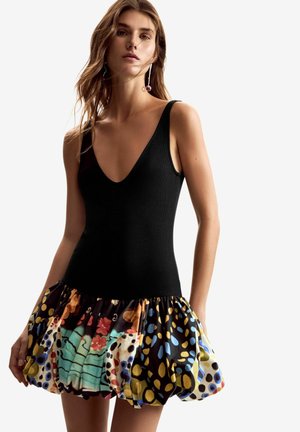 Vrouw draagt een mouwloze zwarte top met een diepe V-hals en een korte, kleurrijke rok met een patroon en geplooide stofdetails.