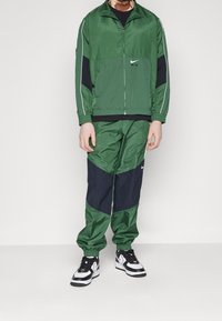 Groene Nike trainingsoutfit met een geassorteerde jas met rits en broek met elastische tailleband. Zwarte accenten en witte strepen. Draagt zwarte en witte sneakers.