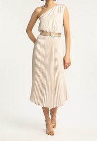 Jednoramienna plisowana sukienka w jasnobeżowej tkaninie, z fakturowanym gorsetem i metalicznym paskiem, zestawiona ze złotymi sandałami na obcasie.