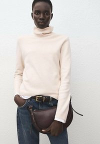 Cremfarbener Rollkragenpullover, blaue Denim-Jeans, braune Ledertasche. Pullover aus weichem Material, Tasche hat eine geschwungene Form und einen Reißverschluss.