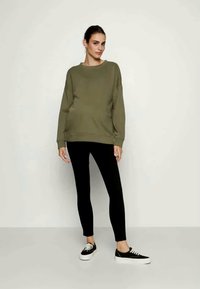 Olivgrüner, langärmeliger Sweatshirt, entspannte Passform, runder Hals, kombiniert mit schwarzen Leggings und schwarzen Turnschuhen, vor einem neutralen Hintergrund.
