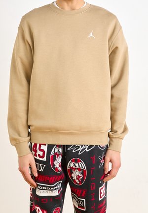Person wearing a beige sweatshirt with a small white logo and black pants with red, white, and gray graphic patches and text.
