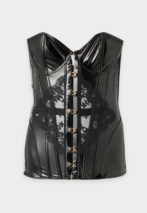 Femme aux longs cheveux foncés portant un corset noir bustier avec des fermetures à l'avant et une lingerie noire assortie, debout pieds nus sur un fond blanc.