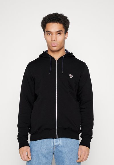 Zwarte zip-up hoodie van zacht materiaal, met een gevoerde capuchon, geribde manchetten en een klein geborduurd logo op de borst. Gecombineerd met lichtblauwe jeans.