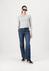 Haut à manches longues gris, jean droit bleu, lunettes de soleil noires, et baskets noires avec des rayures blanches, sur un fond blanc uni.