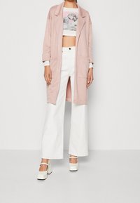 Bledě růžový oversized blazer, bílá zkrácená košile s grafikou, bílé široké kalhoty a bílé boty nablokovém podpatku s řemínkem kolem kotníku.