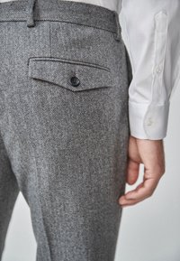 Grijze visgraatbroek met een achterzak met knoopsluiting, rechte voorkant en een soepele tailleband, gedragen met een wit shirt.