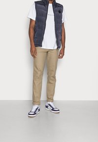 Marinblå pufferväst över en vit T-shirt med texten "MENNAGE", beige byxor och sneakers med marinblå detaljer. Slät textur, avslappnad design.