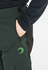 Grüne und schwarze Hose mit einem Reißverschlusstasche, gefertigt aus einem strapazierfähigen Stoff. Enthält ein grünes, rundes Logo auf der Vorderseite und einen Klettverschlussbund.