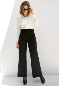 Witte blouse met lange mouwen, ronde halslijn en knoopsluiting, gecombineerd met zwarte wijde broek en spitse zwarte laarzen.