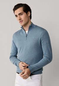 Mladý muž upravujúci rukávy svetlomodrého svetra so štvrťzipsom, oblečený v bielych nohaviciach, stojaci pred jednoducho sivým pozadím.