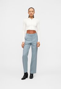 Γυναίκα με πλεξούδες που φοράει λευκή cropped ζακέτα με φερμουάρ, ανοιχτό μπλε ψηλόμεσα παντελόνια και μαύρες μποτάκια στον αστράγαλο, σε λευκό φόντο.