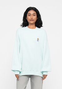 Ilga rankovė, per didelis mėtų spalvos džemperis, pagamintas iš minkštos, tekstūruotos medžiagos, su apvalia iškirpė ir maža grafikine kišenė kairėje krūtinės pusėje.