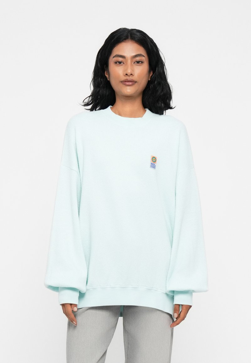 Ilga rankovė, per didelis mėtų spalvos džemperis, pagamintas iš minkštos, tekstūruotos medžiagos, su apvalia iškirpė ir maža grafikine kišenė kairėje krūtinės pusėje.