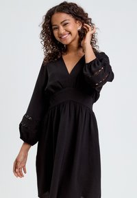 Schwarzes Kleid mit tiefem V-Ausschnitt, langen Puffärmeln mit Ausschnittakzenten und einer taillierten Taille, die in einen ausgestellten Rock übergeht. Weicher Stoff.