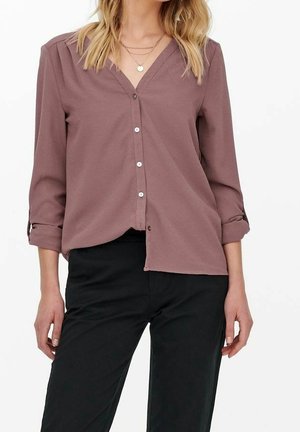 Femme portant un chemisier mauve boutonné avec manches retroussées et pantalon noir, visible du cou jusqu'à la mi-cuisse sur fond blanc.
