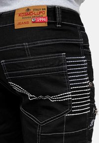 Jeans neri con cuciture bianche a contrasto, più tasche e un'etichetta texturizzata con la scritta "KOSMO-LUPO ROUGH DETAILS."