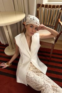 Bela brezrokavna srajca z ostrim ovratnikom in enim gumbom, v kombinaciji s kremnimi čipkastimi hlačami. Model nosi belo kvačkano klobučko in leseno zapestnico.