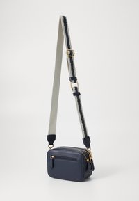 Sac en bandoulière navy avec un extérieur en cuir texturé. Équipé d'une sangle amovible gris clair avec des accents foncés et des éléments en métal doré.