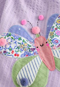 Patch papillon souriant avec des ailes en tissu fleuri et rayé et des antennes pompons roses et violettes sur un fond en tissu lilas texturé.