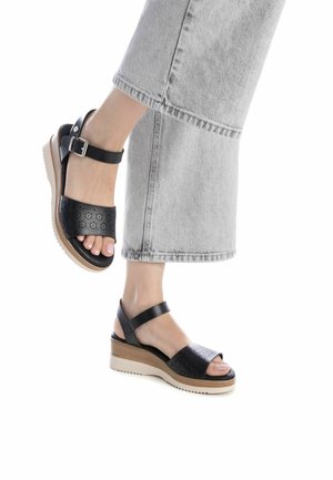 Sandalias de cuero negro perforadas con una correa ancha y hebilla ajustable en el tobillo, que cuentan con una suela de cuña de color claro y un detalle texturizado.