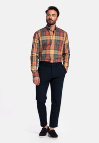 Geruite overhemd in oranje, groen en blauwe tinten, met knoopsluiting en lange mouwen. Gecombineerd met donkere marineblauwe broek en donkere schoenen.