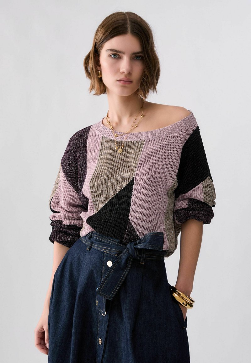 Jeune femme portant un pull en maille multicolore avec des motifs géométriques et une jupe en jean foncé taille haute avec une ceinture nouée.