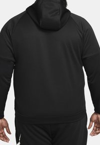 Homme portant une veste à capuche noire unie et un pantalon assorti, vu de dos avec les bras détendus le long du corps.
