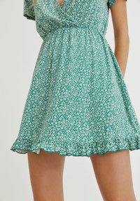 Robe florale verte à manches courtes, taille élastique et ourlet froncé ; présente de petites fleurs blanches et roses sur un fond vert vif.