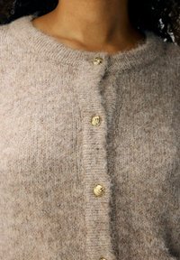 Beige gestrickte Strickjacke mit rundem Ausschnitt und goldfarbenen Knöpfen, die einen strukturierten Wollstoff für Wärme und Komfort präsentiert.