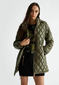 Giacca imbottita di colore verde oliva con trapuntatura, colletto alto, chiusura a zip, vita elastica e dettagli in oro rosa. Indossata sopra un top floreale.